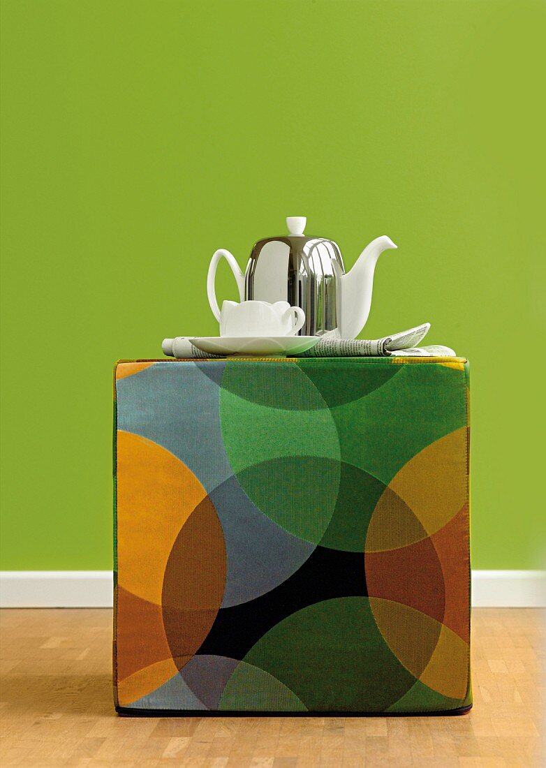 Teekanne und Tasse auf buntem Sitzhocker mit geometrischem Muster im Retro-Stil vor grüner Wand