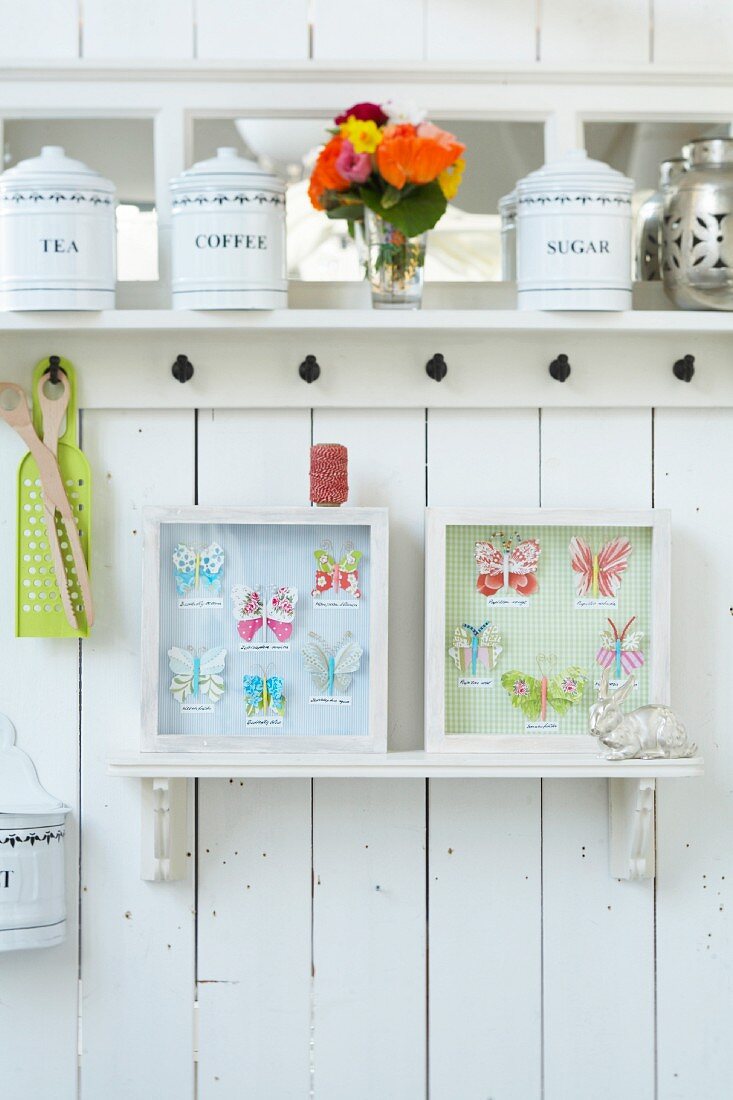Weisses Küchenbord mit alten Aufbewahrungsdosen und Papier-Schmetterlinge in Bildrrahmen an weisser Holzwand