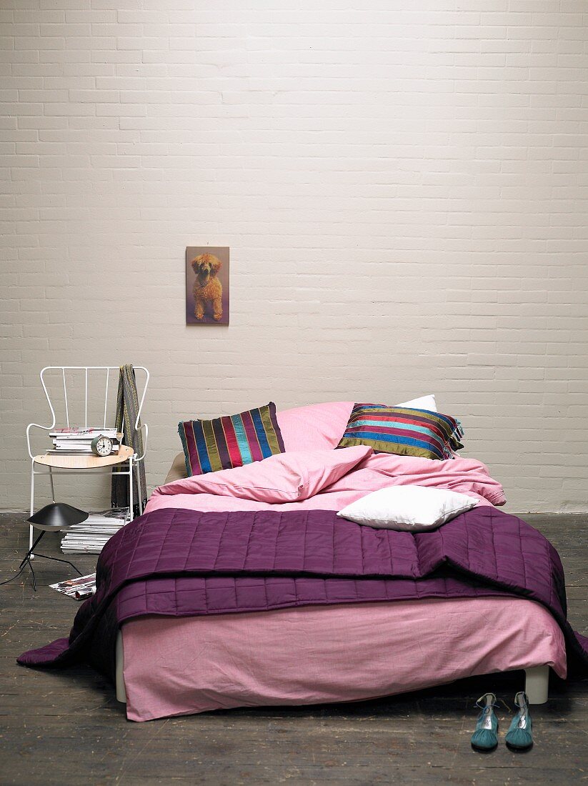 Gemütliches Doppelbett mit lila Plaid und rosa Bettwäsche vor geweisselter Ziegelwand