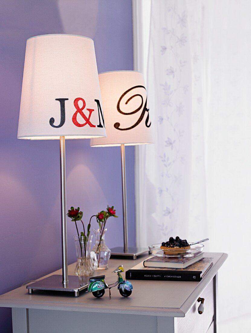 Two table lamps on chest of drawers against lilac wall