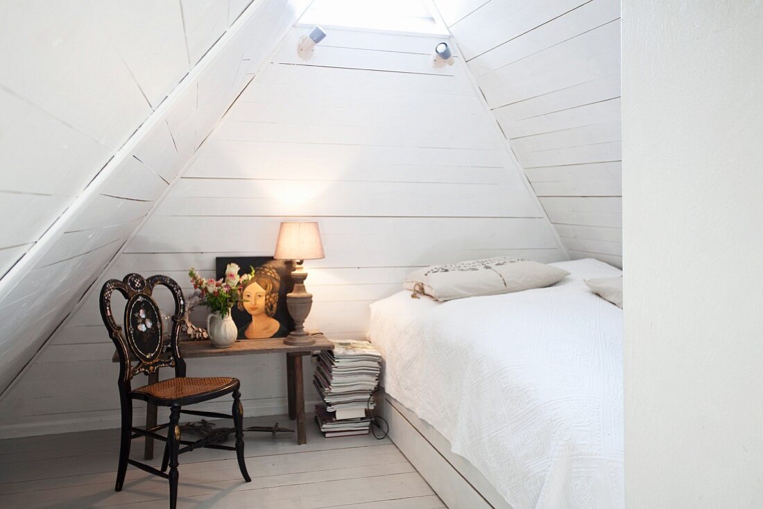 Schlafraum in weiss unter Dachbodengiebel mit Bett, antikem Holzstuhl & Beistelltisch