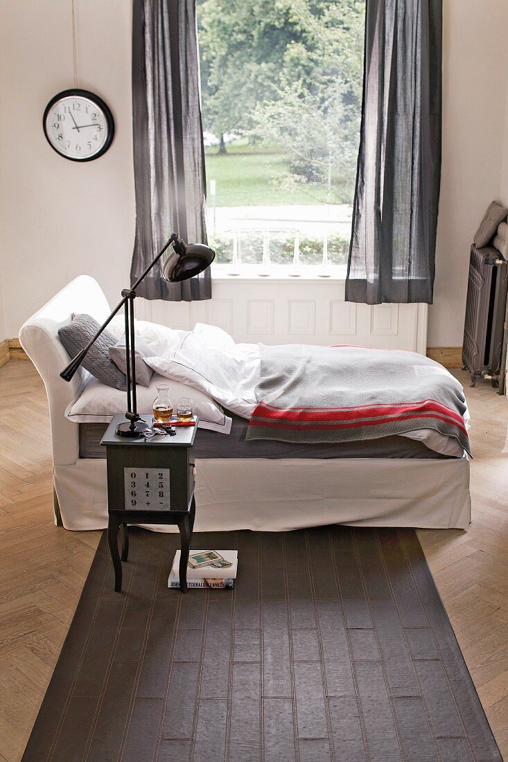 Einzelbett vor Fenster mit Gartenblick, antikes Beistelltischen mit Nachttischleuchte auf grauem Teppichläufer