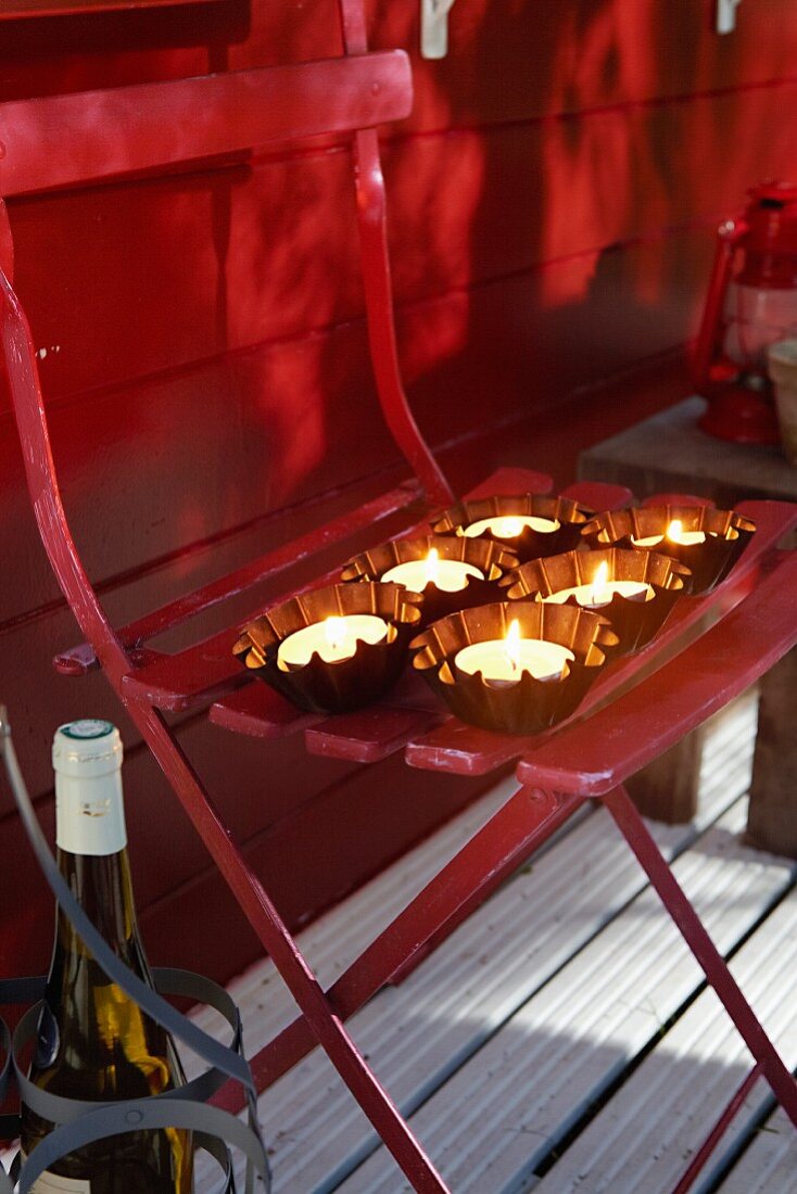 Brennende Teelichter in kleinen Backförmchen auf Klappstuhl im Freien