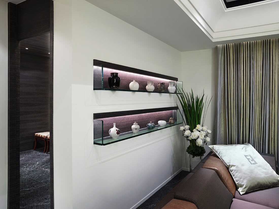 Illuminated niches in wall containing china vases on glass shelves behind sofa in elegant interior