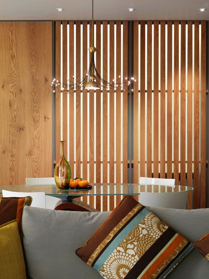 Dekoratives Kissen auf Sofa vor Esstisch mit Glasplatte und Lamellentür aus Holz