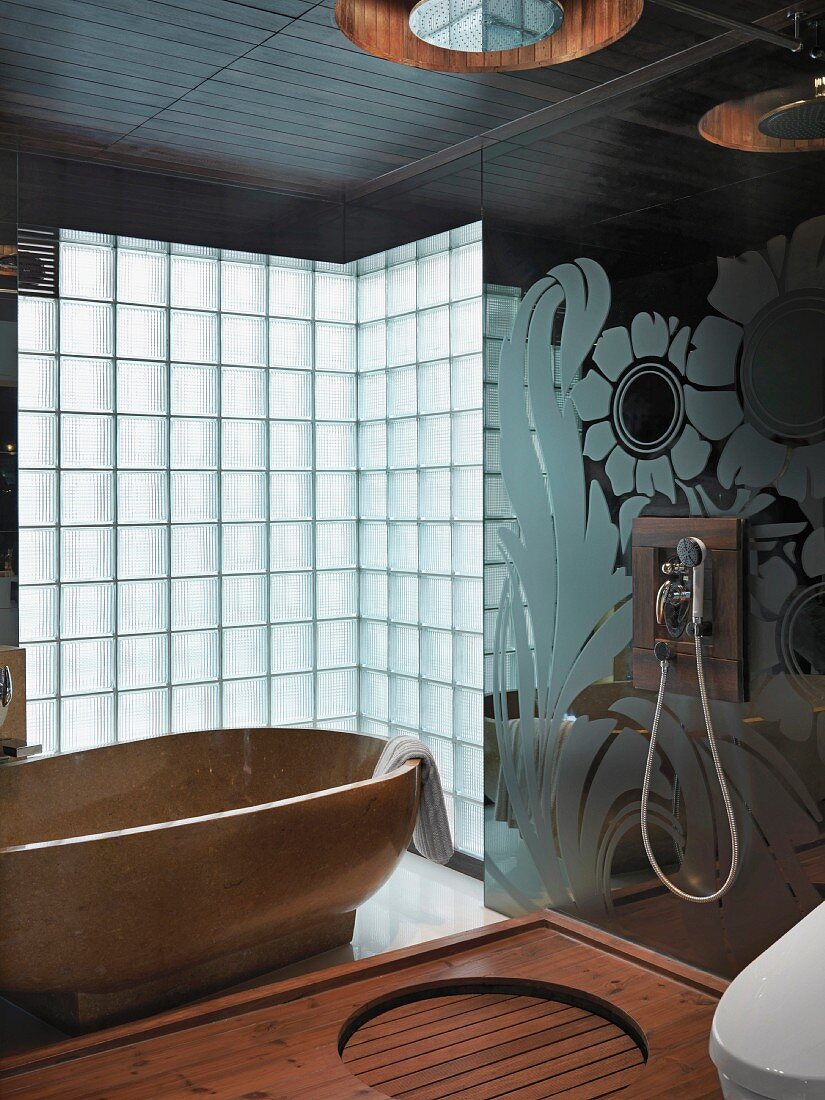 Hand-held shower head on a floral-patterned glass wall and a shower head above a wooden shower area with a stone bathtub in front of glass brick wall in the background