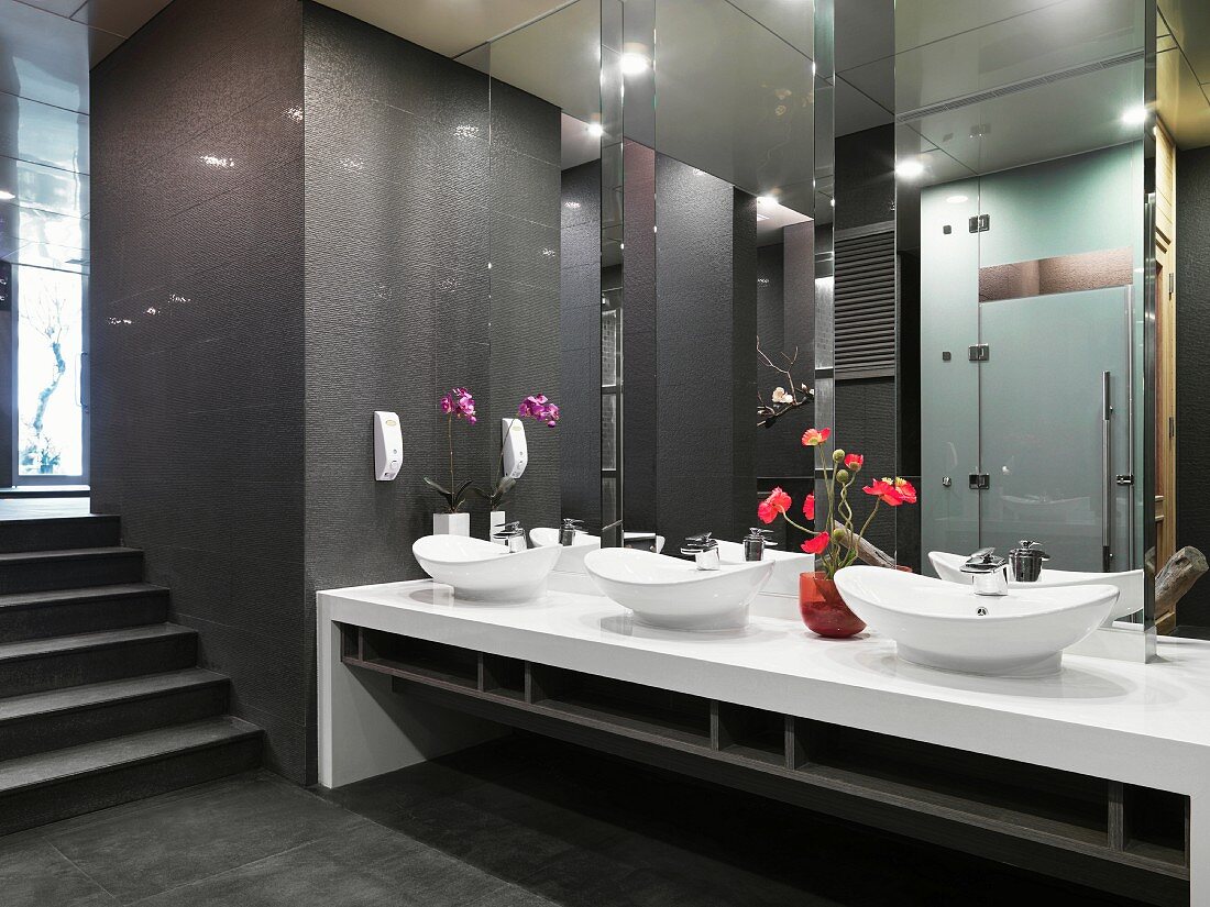 Long mirrored wall above oval washbasins and flower arrangements in Oriental washroom in dark shades