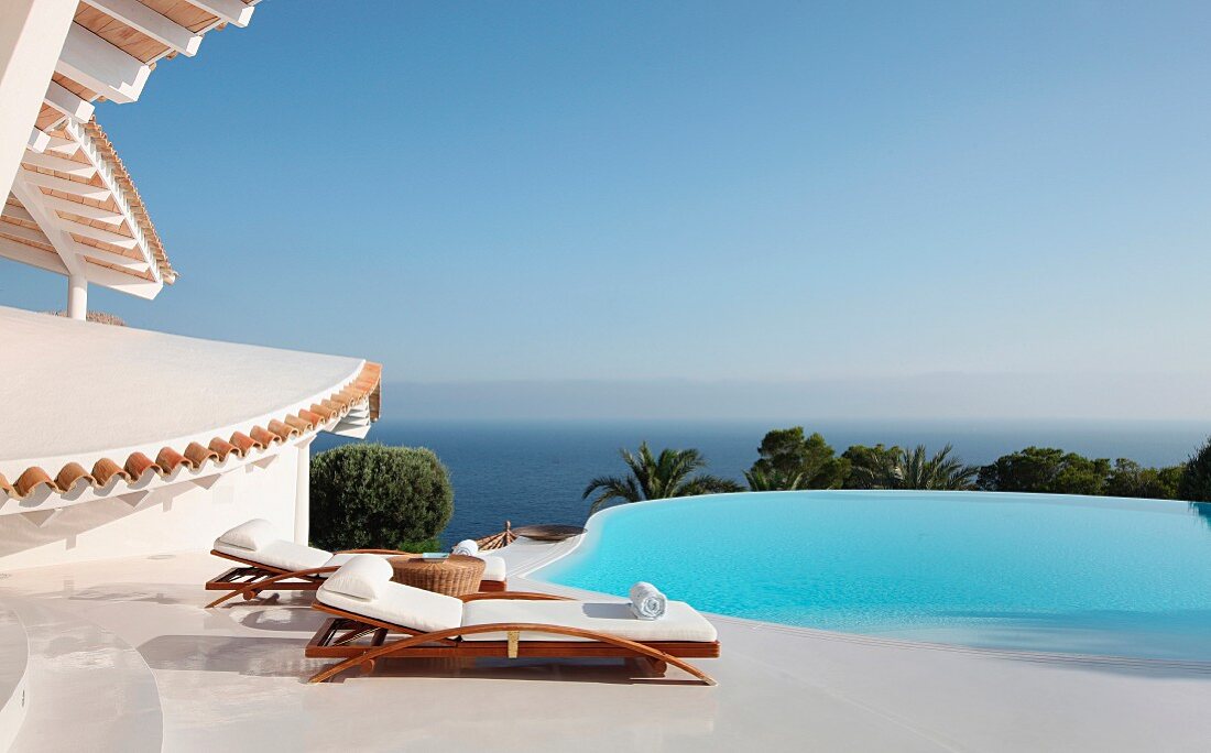 Liegestühle auf weißem Terrassenboden vor Pool mit Meerblick