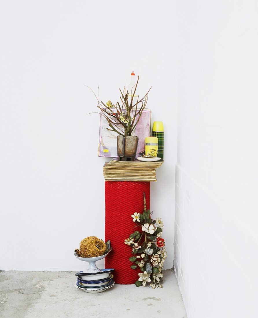Vase of flowering twigs next to storage jars on end of rolled carpet next to vintage metal bowls on concrete floor