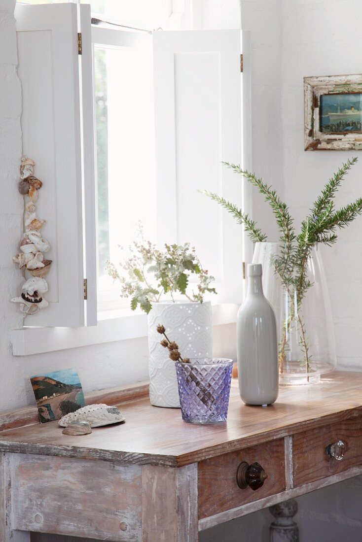 Console table with drawers and weathered white paint below window with interior shutters; various vases and containers of twigs from garden