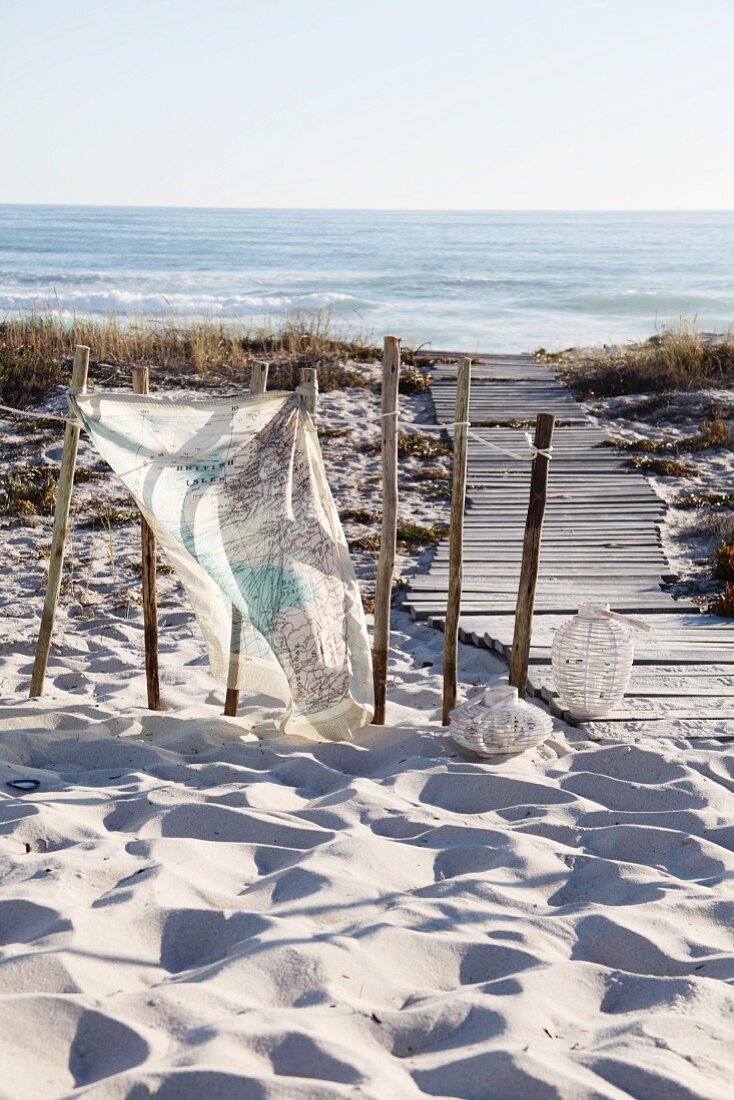 An Holzpflöcke gebundenes, mit Landkartenmotiv bedrucktes Tuch in Sanddünen neben einem Holzsteg zum Meer