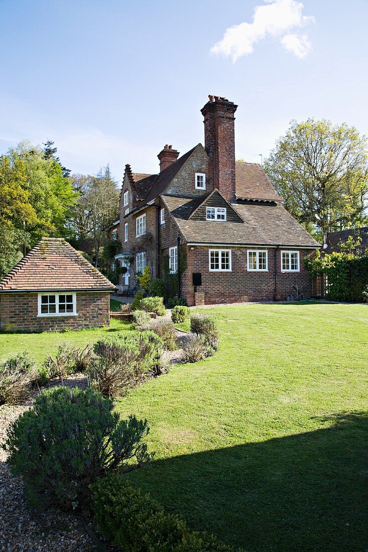 Gepflegter Rasen und gesäumter Kiesweg eines alten, englischen Landhauses mit typisch weissen Sprossenfenstern und Ziegelmauerwerk in sommerlicher Stimmung.
