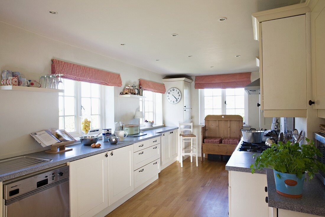Langgestreckte moderne Landhausküche mit alter Holzbank im Hintergrund und rotweiss gestreiften Raffrollos, Parkettboden und integrierter Deckenbeleuchtung.