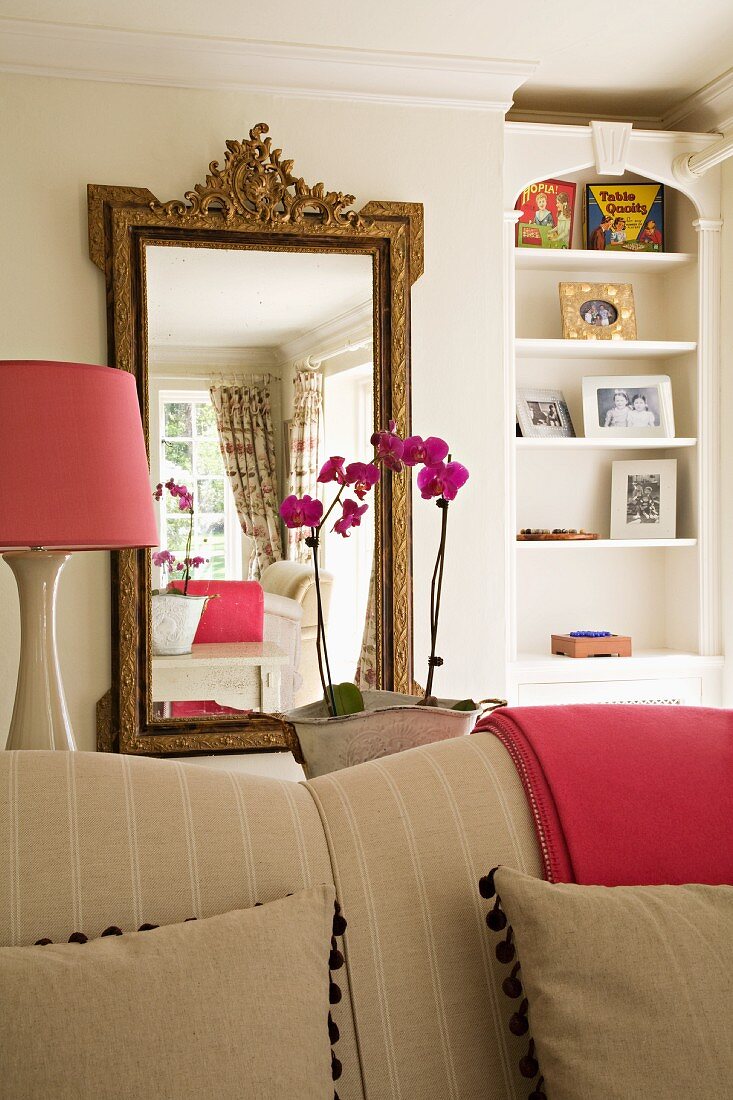 Spiegel mit Goldrahmen im Antikstil hinter beigem Sofa; Lampenschirm und zusammengelegte Decke in sattem Rosa; feminine Stimmung im Wohnbereich