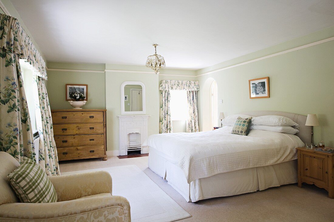 Gepolstertes Doppelbett, rustikale Naturholzkommoden und floral gemusterte Schabrackenvorhänge in großzügigem Landhaus-Schlafzimmer