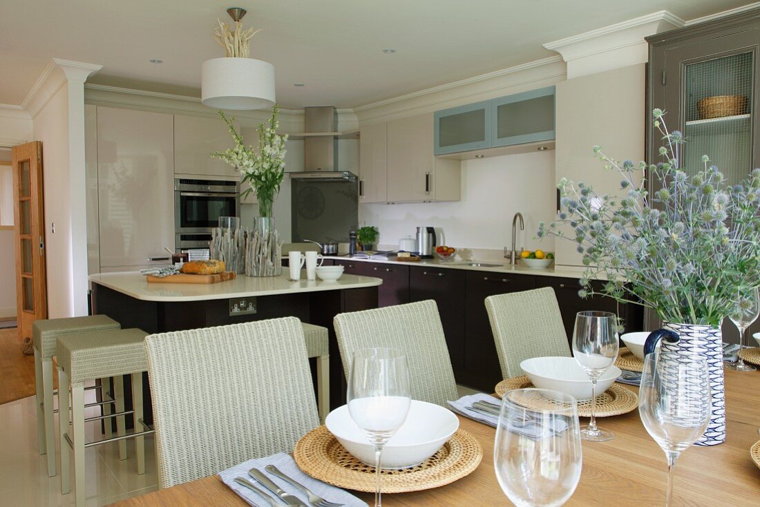 Elegante Küche mit Korbhockern an Küchenblock und festlich gedeckter Tisch mit geflochtenen Platztellern im Vordergrund