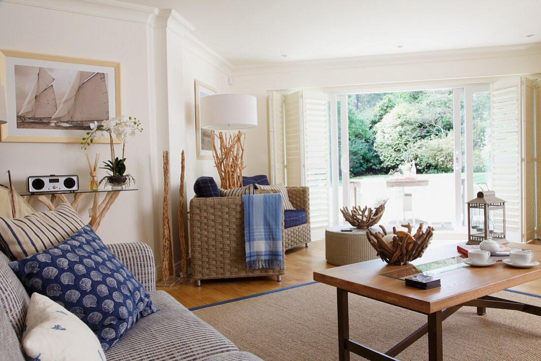 Wicker armchairs, textiles in shades of blue and various accessories made from root wood in open-plan, maritime interior with view of garden