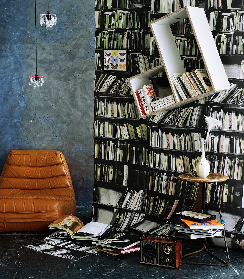 Photographic wallpaper with bookcase motif; slanted shelves mounted on wall & vintage leather chair