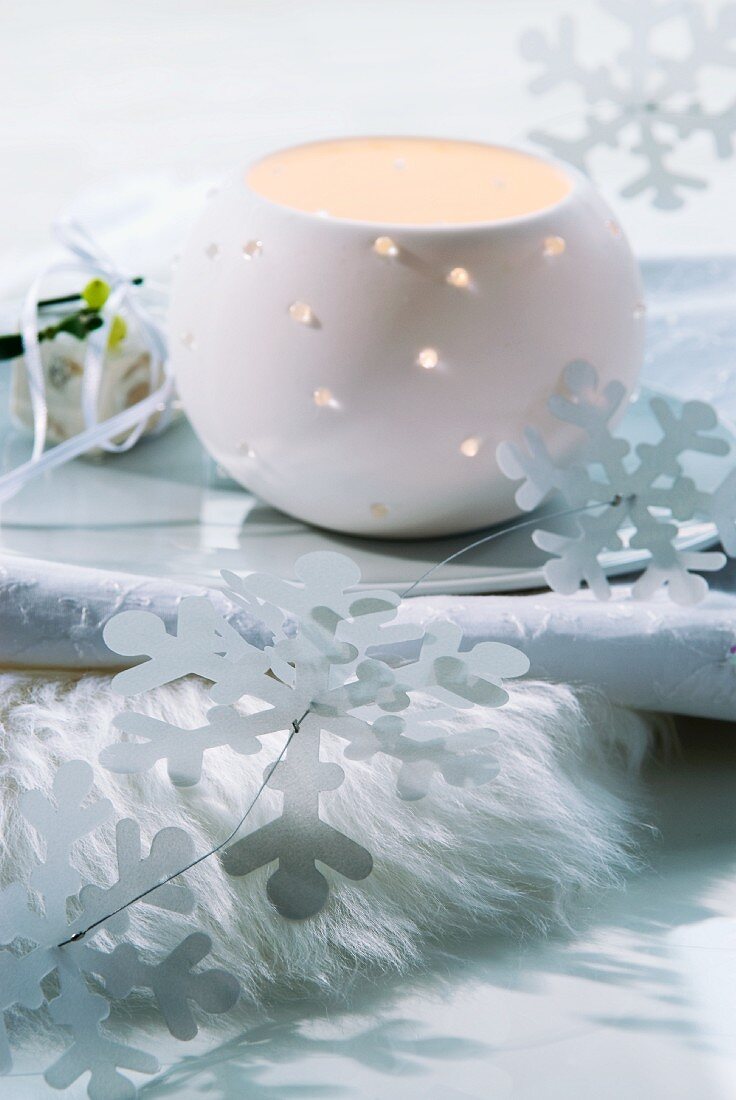 Windlicht aus Porzellan und stilisierte Schneeflocken aus Papier auf Schale und Fellunterlage