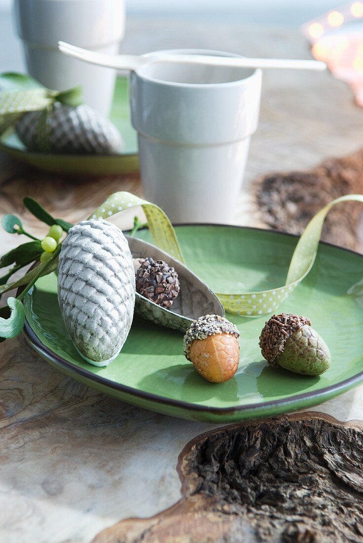 Acorns and fir cones on dish in front of white china beaker and fork on rustic wooden surface