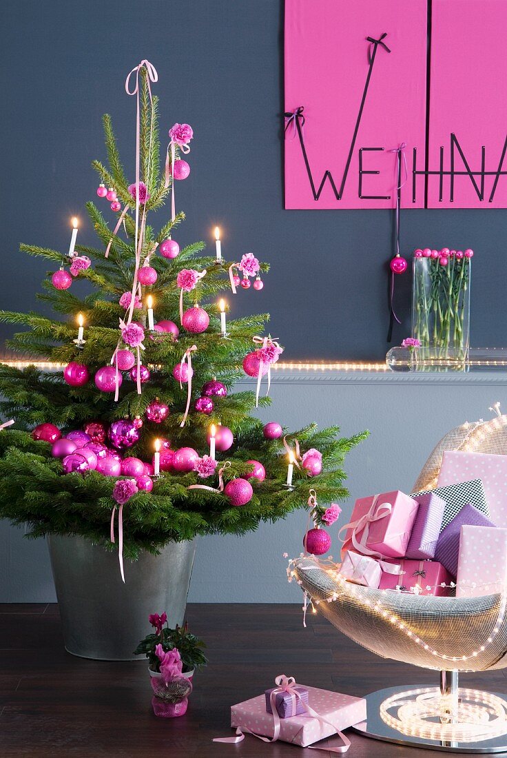 Geschmückter Weihnachtsbaum mit pinkfarbenen Kugeln neben Sessel mit Geschenken vor grauer Wand und Weihnachtsgruss an pinkfabener Tafel