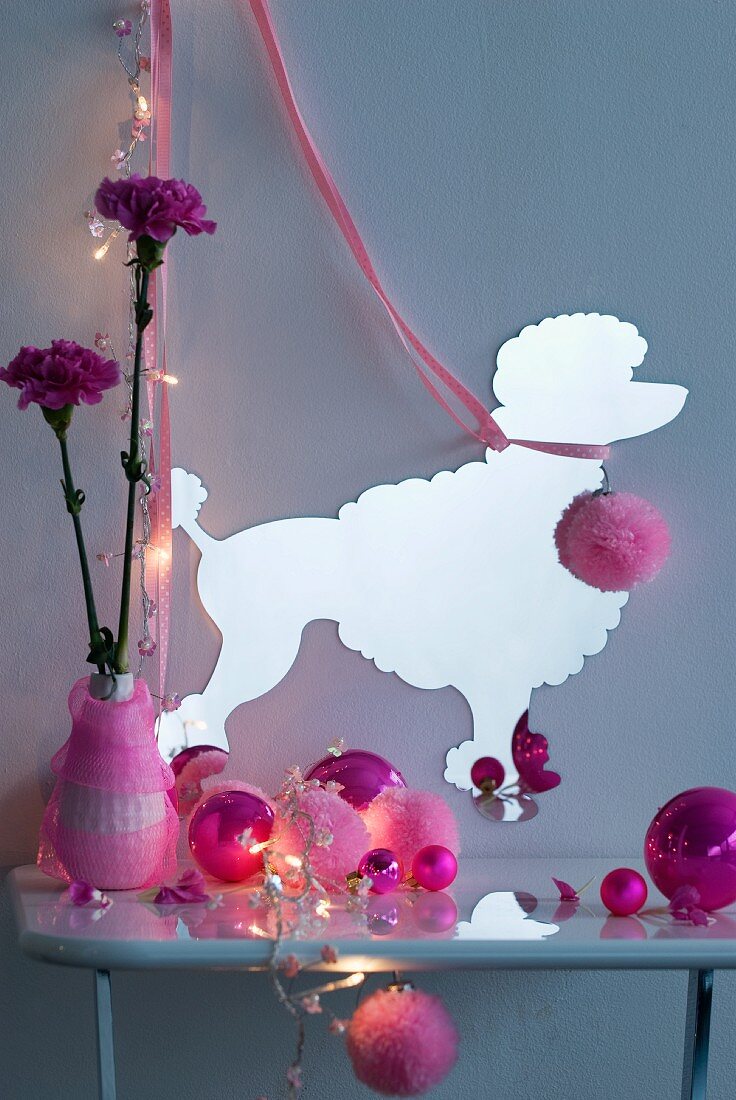Various pink Christmas baubles on console table in front of paper silhouette of poodle on grey wall