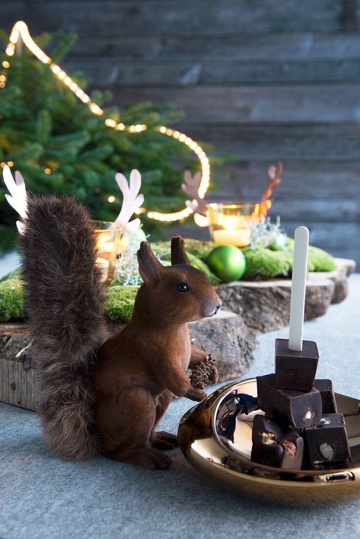 Eichhörnchenfigur vor getürmten Schokostücken in Messingschale und weihnachtlich dekorierter Hintergrund