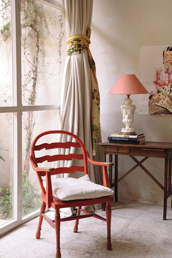 Rustikaler roter Holzstuhl vor Glasfront und Beistelltisch-Sekretär mit traditioneller Tischleuchte in Zimmerecke