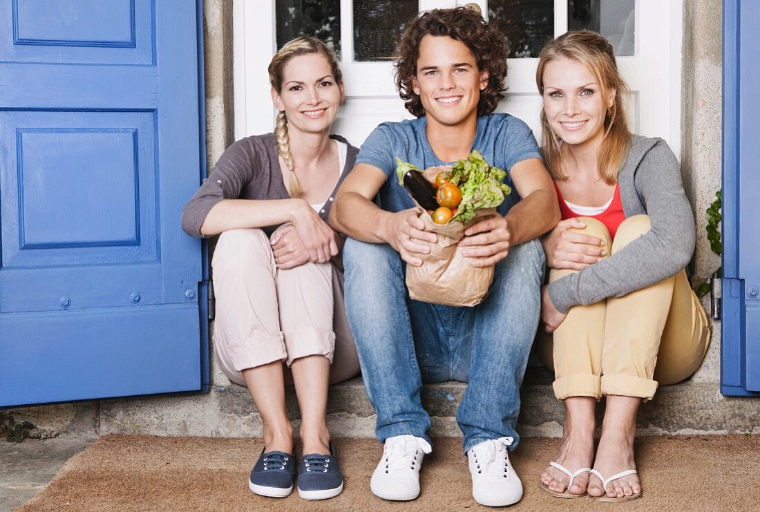 Two young women and a young man sitting in front of a door with vegetables