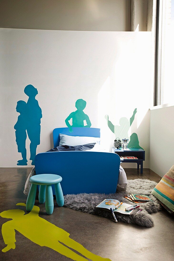 Blue stool in front of single bed with blue frame in corner of room and stencilled silhouettes of people on floor and wall