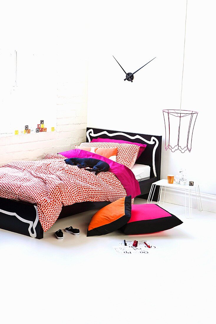 Modern bed with black and white frame and colourful cushions on floor in corner of white room