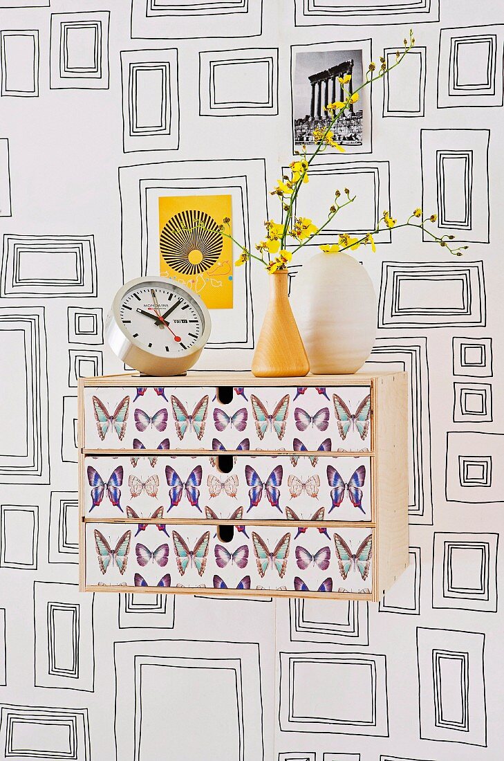 Holzbord mit Schubladen und Schmetterlingsmotiven auf Front vor tapezierter Wand mit Bilderrahmenmotiven