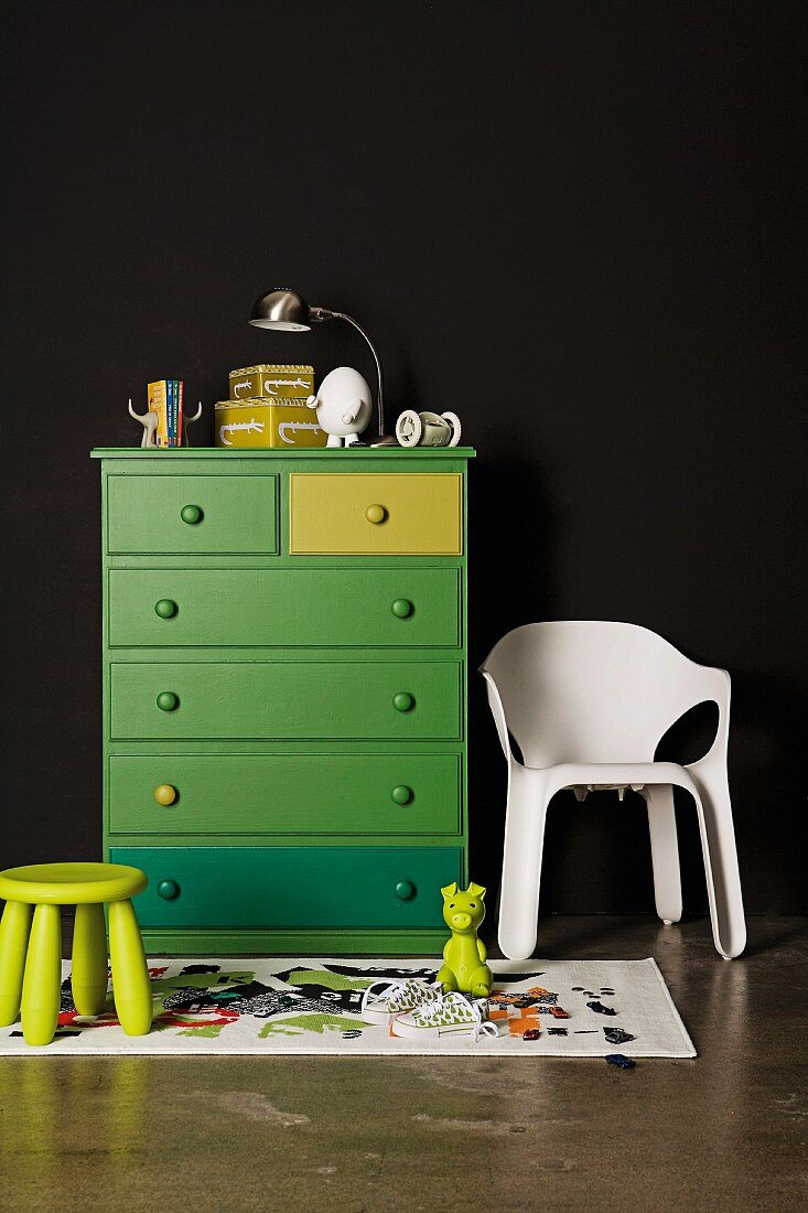 Grüner Hocker vor grün gestrichener Kommode neben weißem Kunststoff Stuhl vor schwarzer Wand