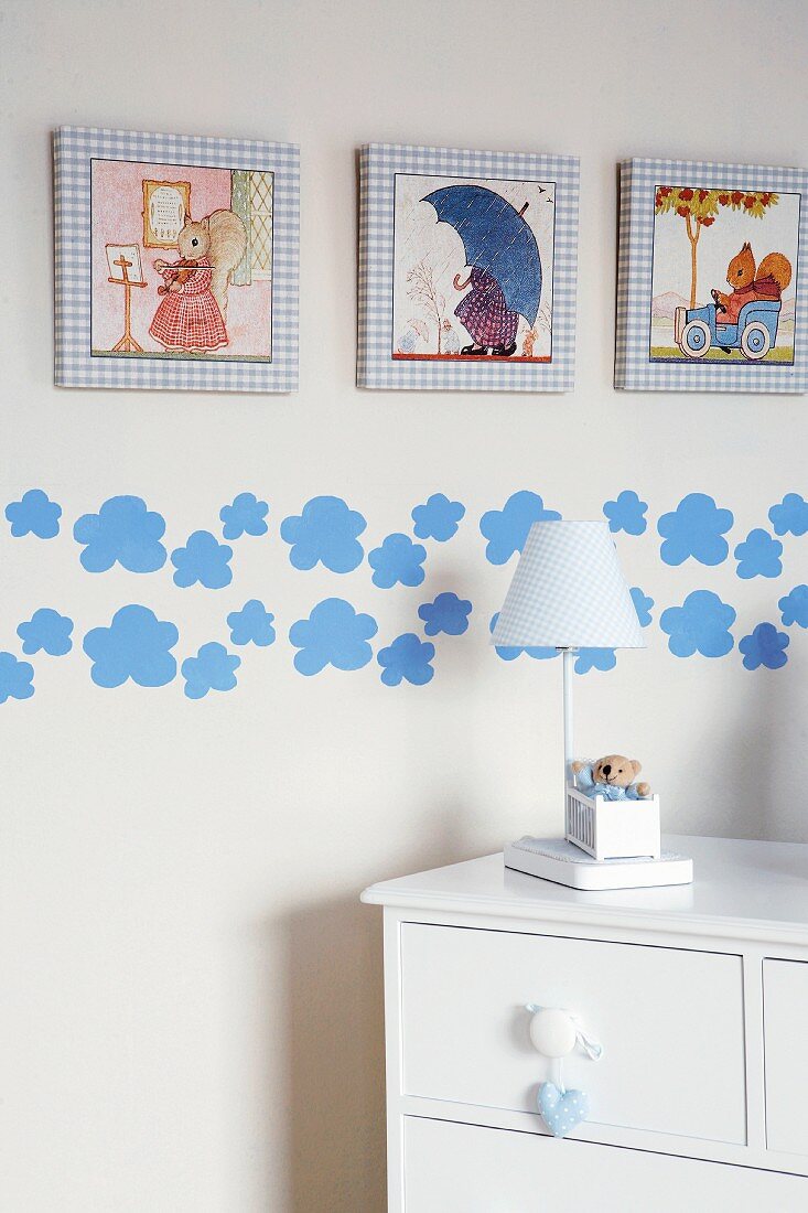 Kinderzimmerausschnitt - weiße halbhohe Kommode mit Tischleuchte vor Wand mit gerahmten Bildern und applizierten Wolkenmotiven