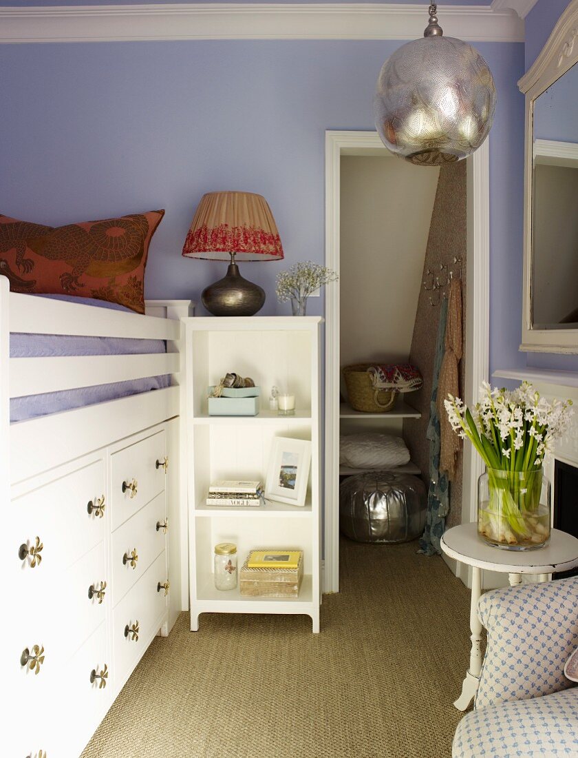 Lilac-painted child's bedroom with raised bed above built-in drawers next to open doorway with shelves in niche