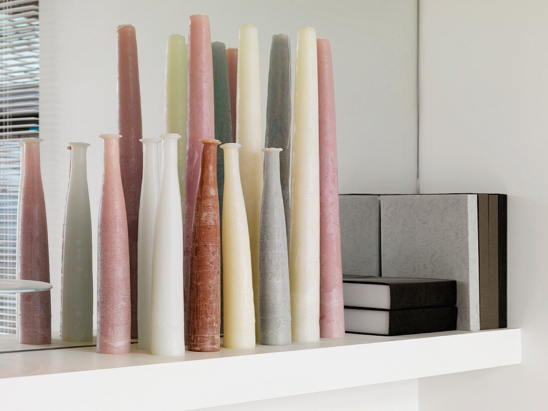 A variety of colored candles next to books on a shelf