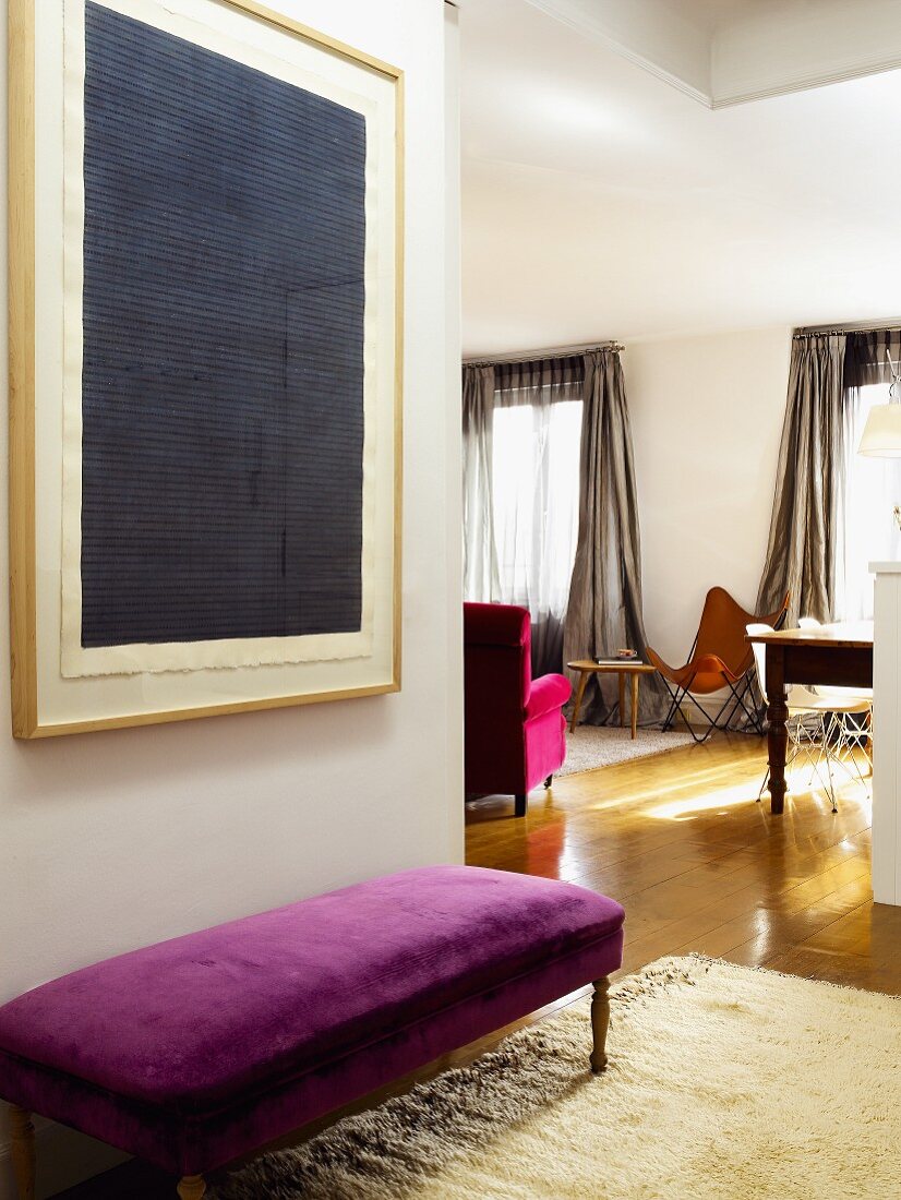 Blick in den offenen Wohnraum; davor eine gepolsterte Sitzbank mit violettem Samtbezug und mit darüberhängendem, modernen Wandbild
