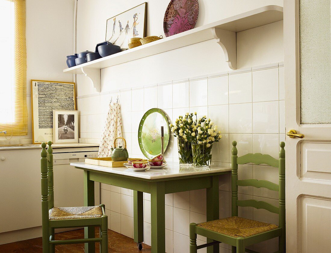 Simple kitchen table and green-painted chairs against tiled wall and below white shelf
