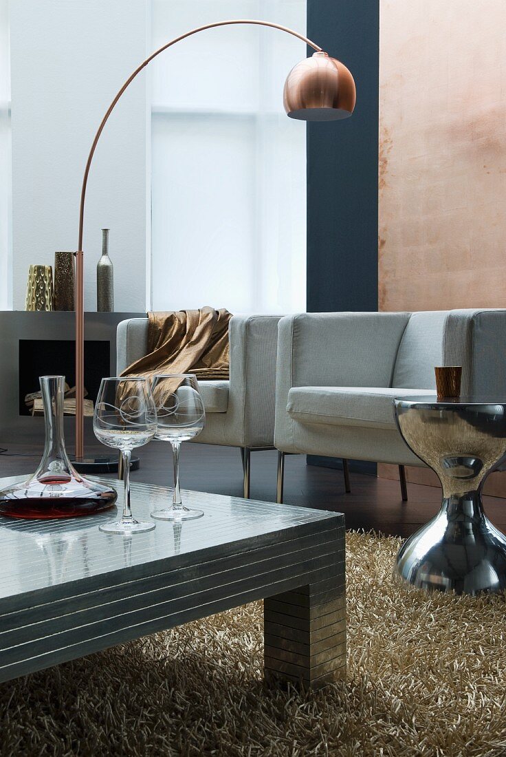 Weinkaraffe und Gläser auf Metall Bodentisch vor modernen Sesseln und kupferfarbener Bogenlampe