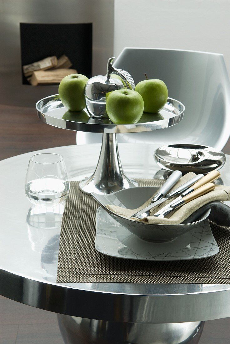Silberschale auf Fuss mit grünen Äpfeln und Schale mit Besteck auf rundem Tisch