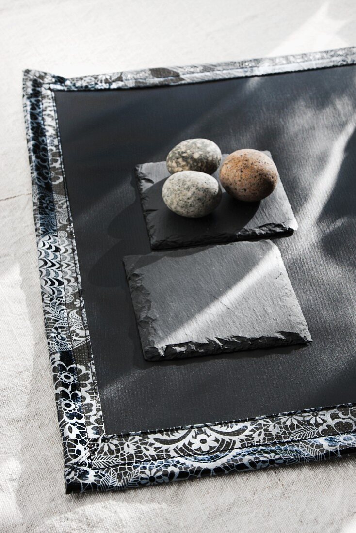 Drei Kieselsteine auf Schieferplatte und schwarzem Tischset mit gemusterter Einfassung