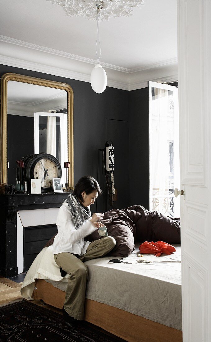 Lady sitting on a bed sewing in a bedroom with dark gray walls, in the background a gold framed mirror and clock over the fireplace