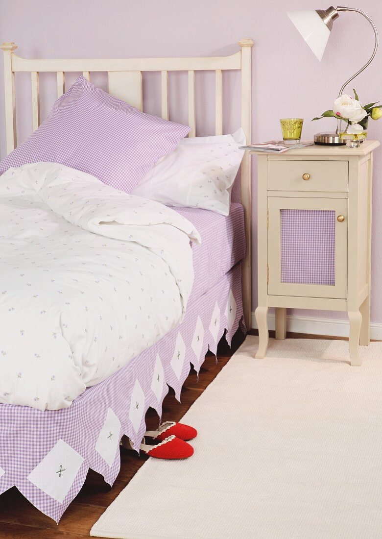 Country-style single bed with white, wooden, slatted headboard next to bedside cabinet with table lamp against lilac wall