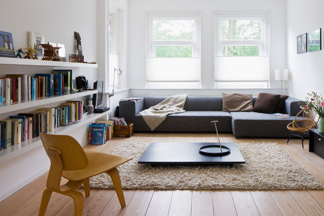 Niedriger Bauhaus Stuhl aus Holz vor dunklem Bodentisch auf flokatiartigem Teppich und graues Designer Sofa am Fenster