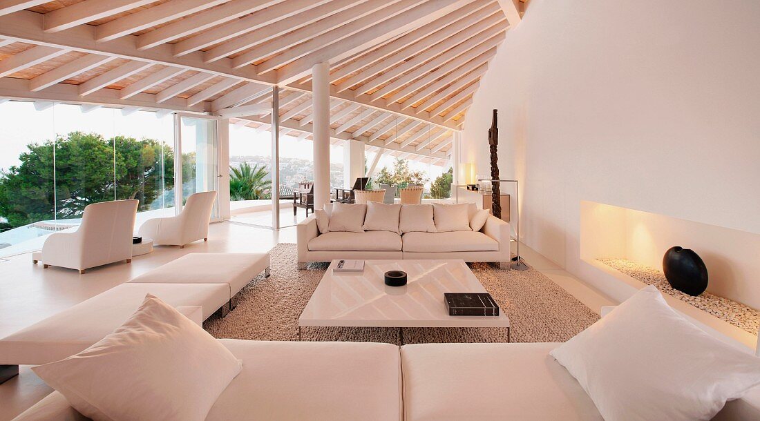 Sofas und Hocker gruppieren sich um einen minimalistischen Couchtisch in einem weissen Wohnzimmer mit Holzbalkendecke und Panoramafensterfront
