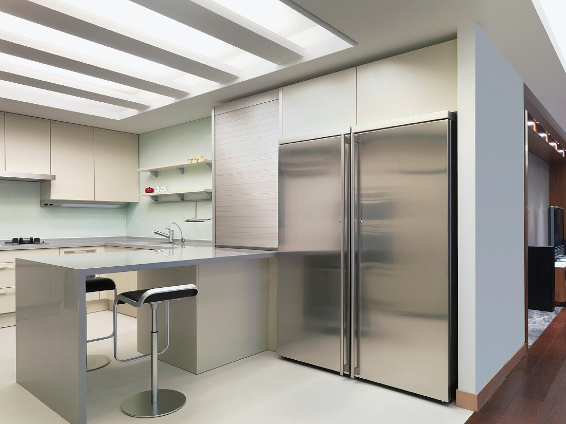 Large stainless steel refrigerator in a modern built-in kitchen with a breakfast bar and indirect ceiling lighting