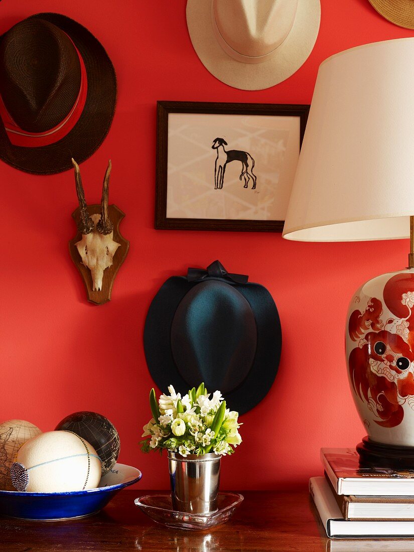 Kleiner Blumenstrauss im Metallbecher zwischen Tischlampe und Teller mit Dekofrüchten vor rotgetönter Wand und aufgehängter Hutsammlung