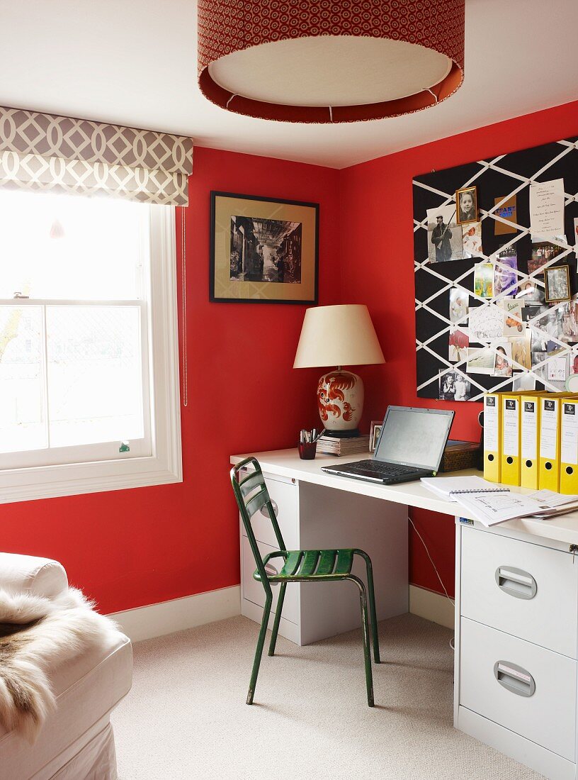 Grüner Metallstuhl vor hellem Schreibtisch an rot getönter Wand und Fenster mit gemustertem Rollo