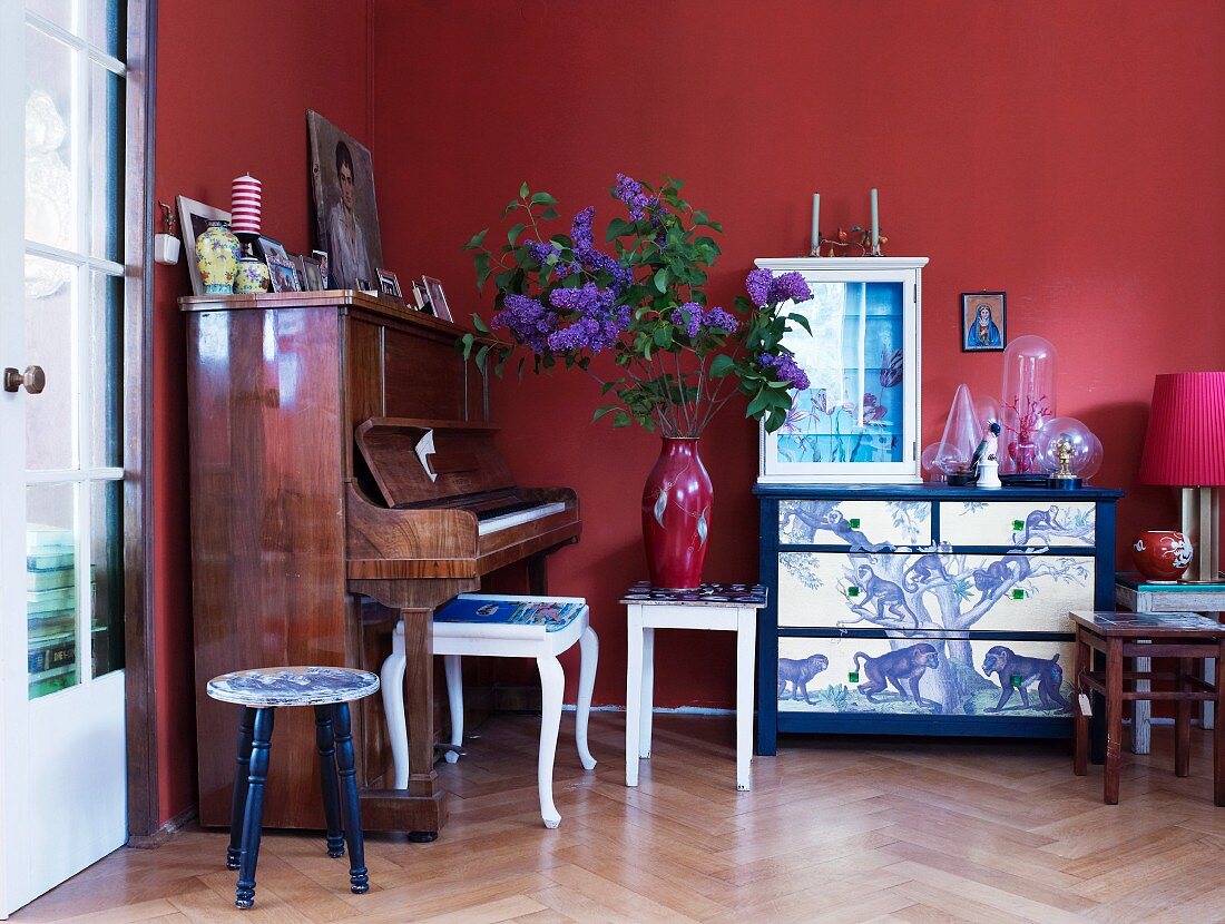 Glasvitrine auf kunstvoll bemalter Kommode, ein altes Klavier und verschiedene Vintage-Hocker vor rot getönter Wand
