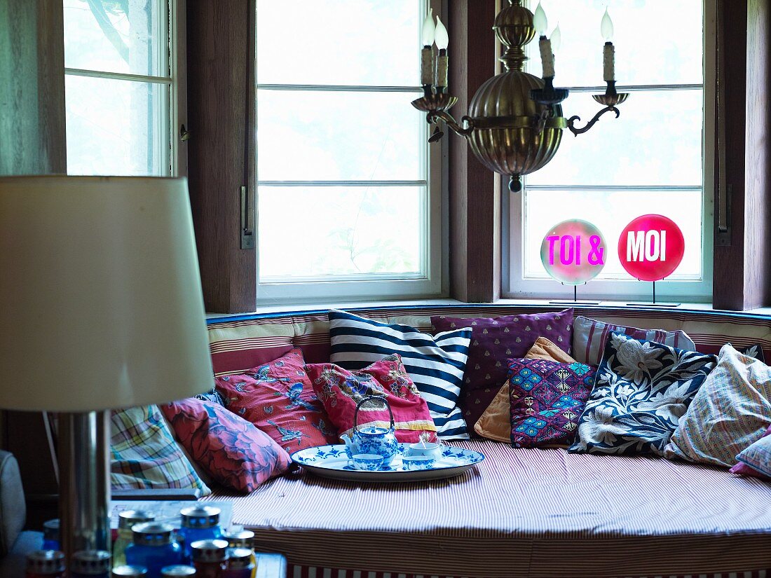 Einladendes Sofa mit vielen Kissen und Teetablett in halbrundem Fenstererker mit leuchtenden TOI & MOI-Schildern
